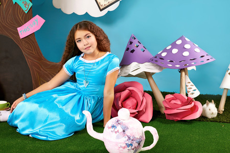 Little beautiful girl with long wavy hair in a blue silk dress in the scenery of Alice in Wonderland with white fluffy bunny cartoon fairy tale adventureの写真素材