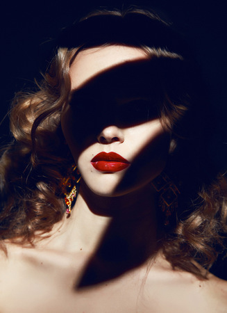Beautiful young sexy blonde girl with curly hair with bright makeup red lips, red lipstick, long earrings in dark shadowsの写真素材