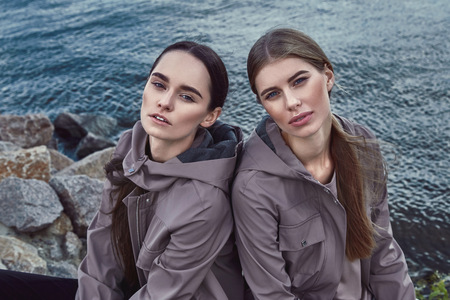 Two beautiful sexy woman walk on beach water wear jacket suit accessory fashion style autumn casual collection natural organic clothes long blonde brunette hair ocean sea beach view water cloud skyの写真素材