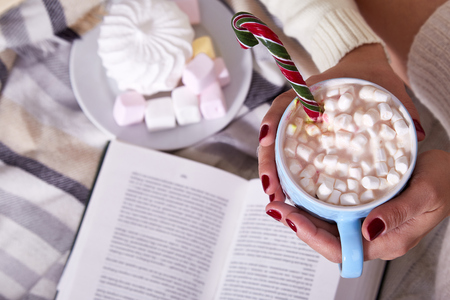 Beautiful Merry Christmas morning woman drink cup sweet hot cacao with marshmallow eat lollipop candy cane wear home knitted wool plaid laptop New Year eve read book winter mood manicure nail polish.の写真素材