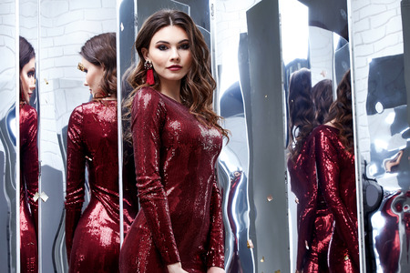 Beautiful sexy lady pretty face make-up cosmetic long brunette hairstyle beauty salon jewelry earrings woman wear skinny shiny sequins red dress party style fashion clothes for evening celebration.の写真素材
