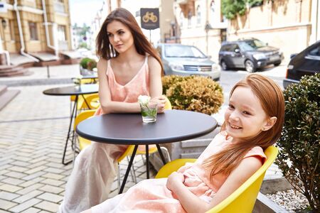 Mother and daughter sitting in street cafe summer time happy family day beautiful cute woman and small little girl in restaurant breakfast holiday morning baby mom buildings fashion dress look.の写真素材