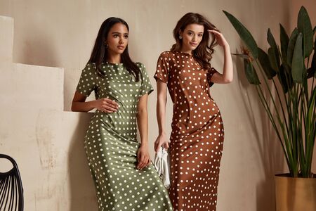 Two fashion model brunette hair wear green  brown dots silk dress sandals shoes accessory clothes date party walk interior journey summer collection plant flowerpot stairs beautiful woman tan skin friends.の写真素材