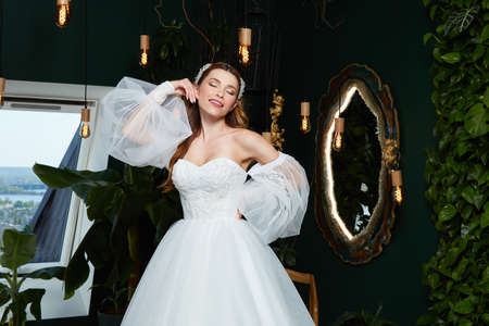 Beautiful tender young woman sexy brunette bride in a luxury white wedding dress lace chiffon Summer happiness awaits the groom before the wedding ceremony makeup accessory jewelry smile.の写真素材