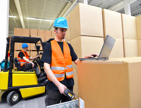 worker with laptop and barcode scanner checking a shipment in the department store - forklift truck drivers in the backgroundの写真素材