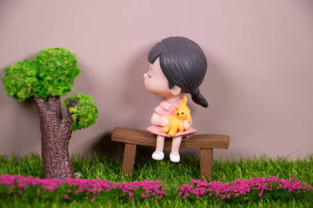 Cute miniature girl figurine sitting on a wooden bench holding a yellow bunny doll, small tree and flowers, pastel background with copy space.の写真素材