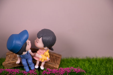 Adorable miniature children figurines holding hands on a wooden bench, sharing a sweet moment with a toy, surrounded by green grass and pink flowers with copy space.の写真素材