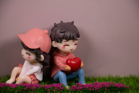 Cute miniature couple figurines sitting back to back on grass, boy holding a red heart, pastel background with copy space, love concept.の写真素材