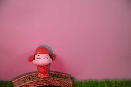 Cute boy figurine in a red traditional outfit standing on a wooden bridge with a pink background and copy space, expressing happiness and cultural charm.の写真素材