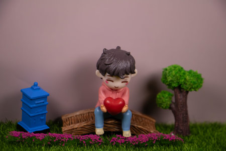 Cute miniature boy figurine sitting on a wooden bridge holding a red heart, small tree and blue...