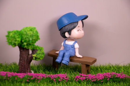 Cute miniature boy figurine wearing blue overalls and cap, sitting on a wooden bench in a small garden with tree and flowers, pastel background.の写真素材