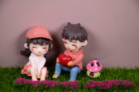 Adorable miniature couple sitting on green grass, a boy holding a red heart beside a smiling girl, with a cute mushroom house and pink flowers on a soft gray background.の写真素材