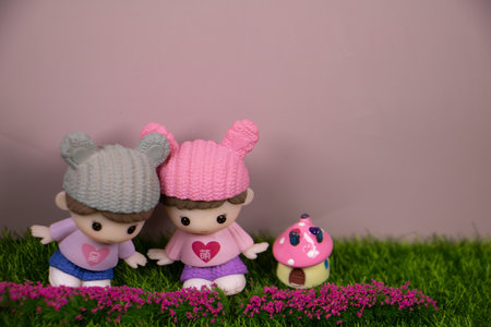 Cute miniature twin dolls wearing knit hats, standing on green grass beside a tiny mushroom house, with pink flowers and a soft pastel background.の写真素材