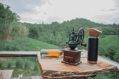 Vintage manual coffee grinder and travel thermos on wooden table with green mountain landscape background. Outdoor coffee setup symbolizing slow life, relaxation, camping and nature lifestyle.の写真素材