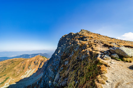 A wide panoramic view of a rocky hiking trail winding through the slopes of the Low Tatras National Park during a sunny autumn day. Beautiful yellow grass and blue skyの写真素材