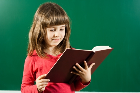 Cute little girl is standing in front of blackboard and reading a bookの写真素材