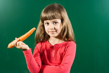 Cute little girl holding carrot in front of blackboard の写真素材