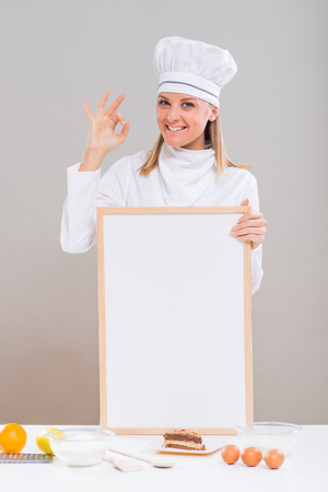 Beautiful female confectioner is showing ok sign while holding whiteboard with slice of cake and ingredients for cake at the tableの写真素材