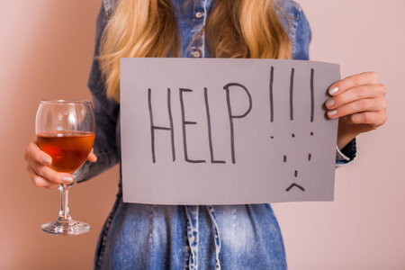 Woman holding alcohol and  paper with word help while standing in front of wall.