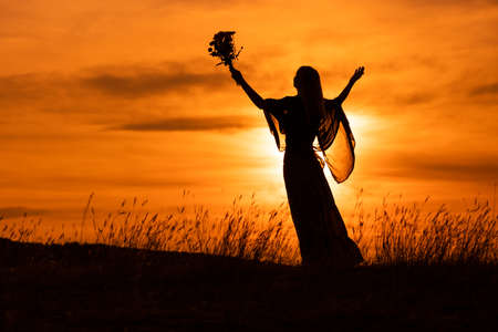 Silhouette of a woman with arms outstretched holding flowers and  looking at beautiful sunset.の写真素材