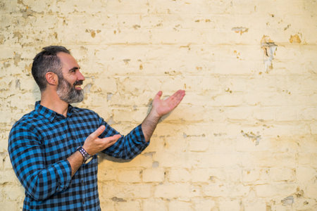 Portrait of modern businessman with beard gesturing while standing in front of brick wall outdoor.
