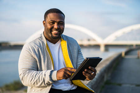 Modern black man using digital tablet while sitting by a river.の写真素材