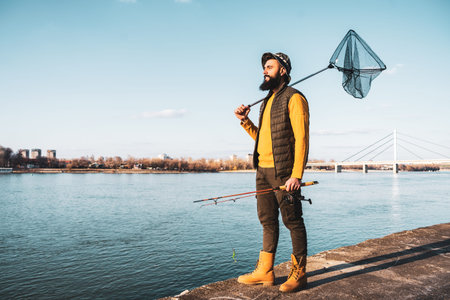 Image of fisherman holding fishing rod  and fishing net while standing by a river.
