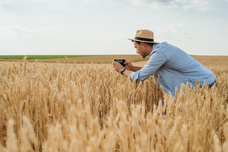 Happy farmer photographing crops with phone while standing in his growing wheat field.