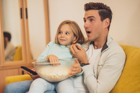 Surprised father and daughter watching tv and eating pop corns together at their home.の写真素材