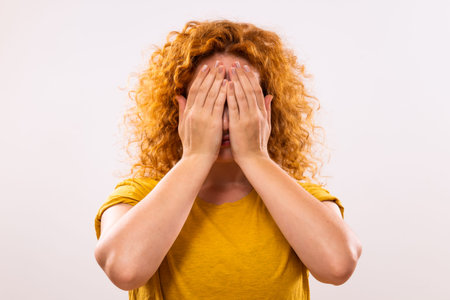 Image of desperate ginger woman covering her face on gray background.