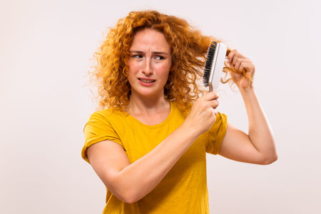 Angry ginger woman holding hairbrush and combing her curly  hair.