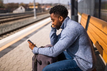 Angry man with a suitcase and mobile phone sitting on a bench at the railway station.の写真素材