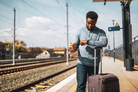 Tired man looking at clock and drinking coffee while standing with suitcase on a railway station.の写真素材