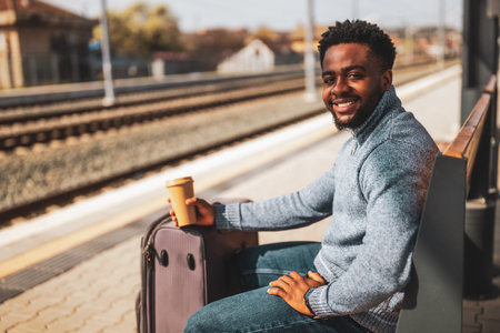 Happy man with suitcase enjoys drinking coffee while sitting on a bench at the railway station.の写真素材