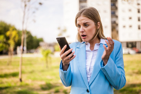 Image of shocked businesswoman looking at her mobile phone while standing outdoor.の写真素材