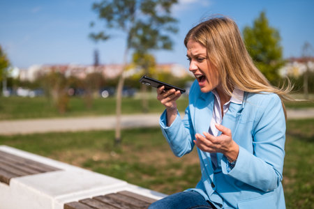 Image of angry businesswoman screaming on the phone while sitting on the bench outdoor.