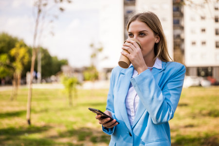 Beautiful businesswoman drinking coffee and using mobile phone while standing outdoor on a sunny day.の写真素材