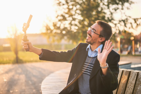Businessman vlogger in coat waving while using mobile phone with gimbal. He is sitting on a bench in the park and recording vlog.の写真素材