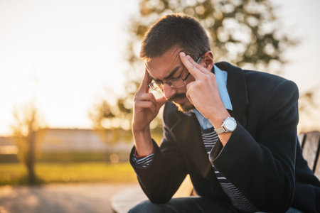 Tired businessman in coat with eyeglasses having a headache while sitting on the bench in park on a sunny autumn afternoon.の写真素材