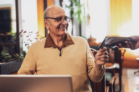 Happy senior man with eyeglasses in casual clothing making a contactless payment with credit card while sitting  in the cozy city cafe at evening.の写真素材