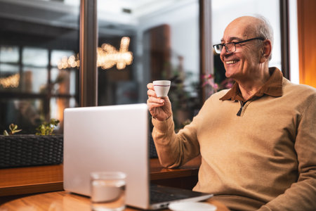 Smiling senior man with eyeglasses using laptop and drinking coffee while sitting in the city cafe at evening.の写真素材