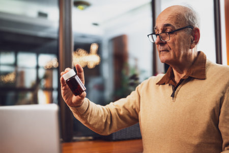 Senior man with eyeglasses holding bottle of pill and using laptop while sitting in the cafe.の写真素材