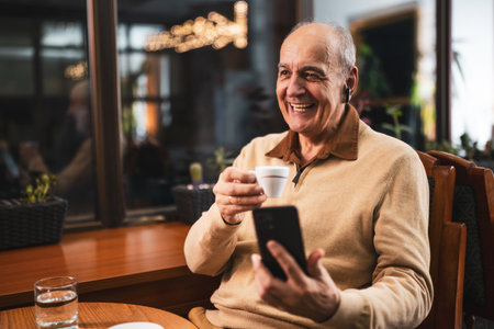Happy senior man drinking coffee while using mobile phone and earphones. He is relaxing in the cozy city bar at evening and using technology.の写真素材