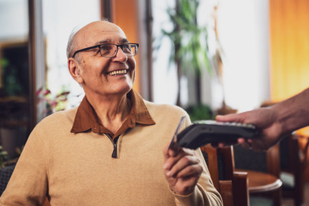 Happy senior man with eyeglasses in casual clothing making a contactless payment with credit card while sitting  in the cozy city cafe at evening.の写真素材