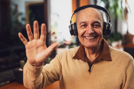 Portrait of senior man with wireless headphones looking at camera and waving during a video call on his laptop.の写真素材