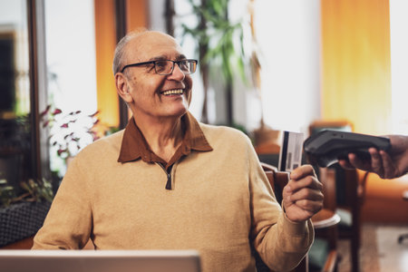 Smiling senior man with eyeglasses in casual clothing making a contactless payment with credit card while sitting  in the cozy city cafe at evening.の写真素材
