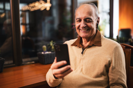 Happy senior man in casual clothing using smart phone and earphones while sitting in the city bar at evening.の写真素材