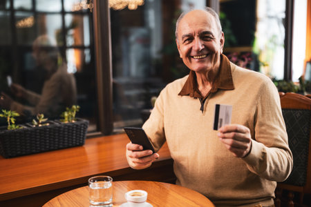 Smiling senior man in casual clothing using mobile phone and credit card while enjoying drinking coffee in the cozy city cafe.の写真素材