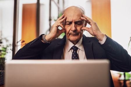 Close up photo of overworked senior businessman in suit having a headache while working on laptop in...