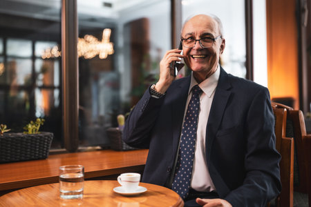 Senior businessman with eyeglasses in elegant suit talking on mobile phone wile enjoying drinking coffee and sitting in the cozy city cafe.の写真素材